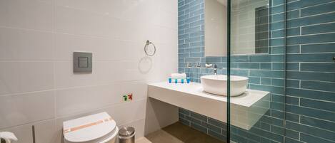 A modern bathroom with a white toilet, a trash bin, a glass shower enclosure, and a sink with a blue-tiled backsplash.