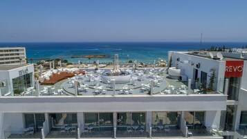 A rooftop terrace with white tables and chairs, a central fountain, and a view of the sea and beach.