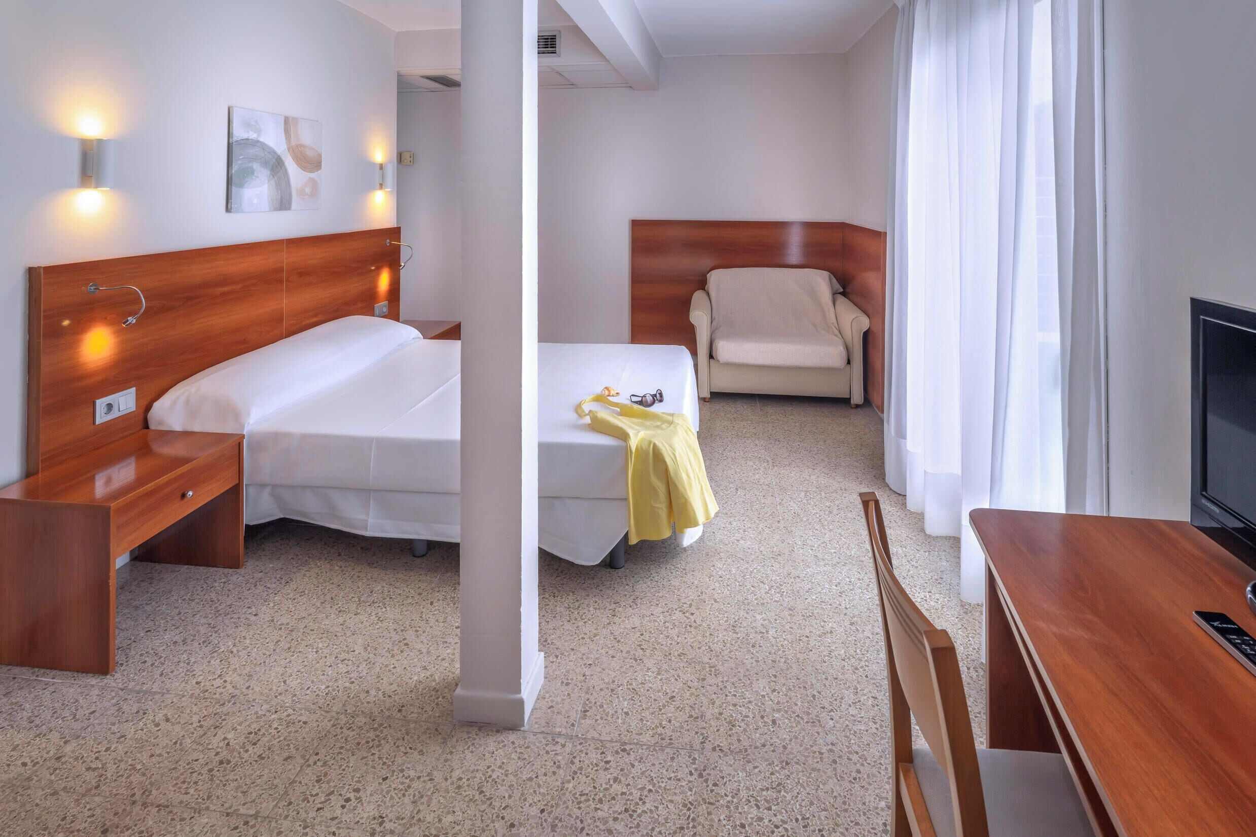 Habitación de hotel con cama de cabecera de madera, mesita de noche, escritorio pequeño con silla, sofá y televisor.