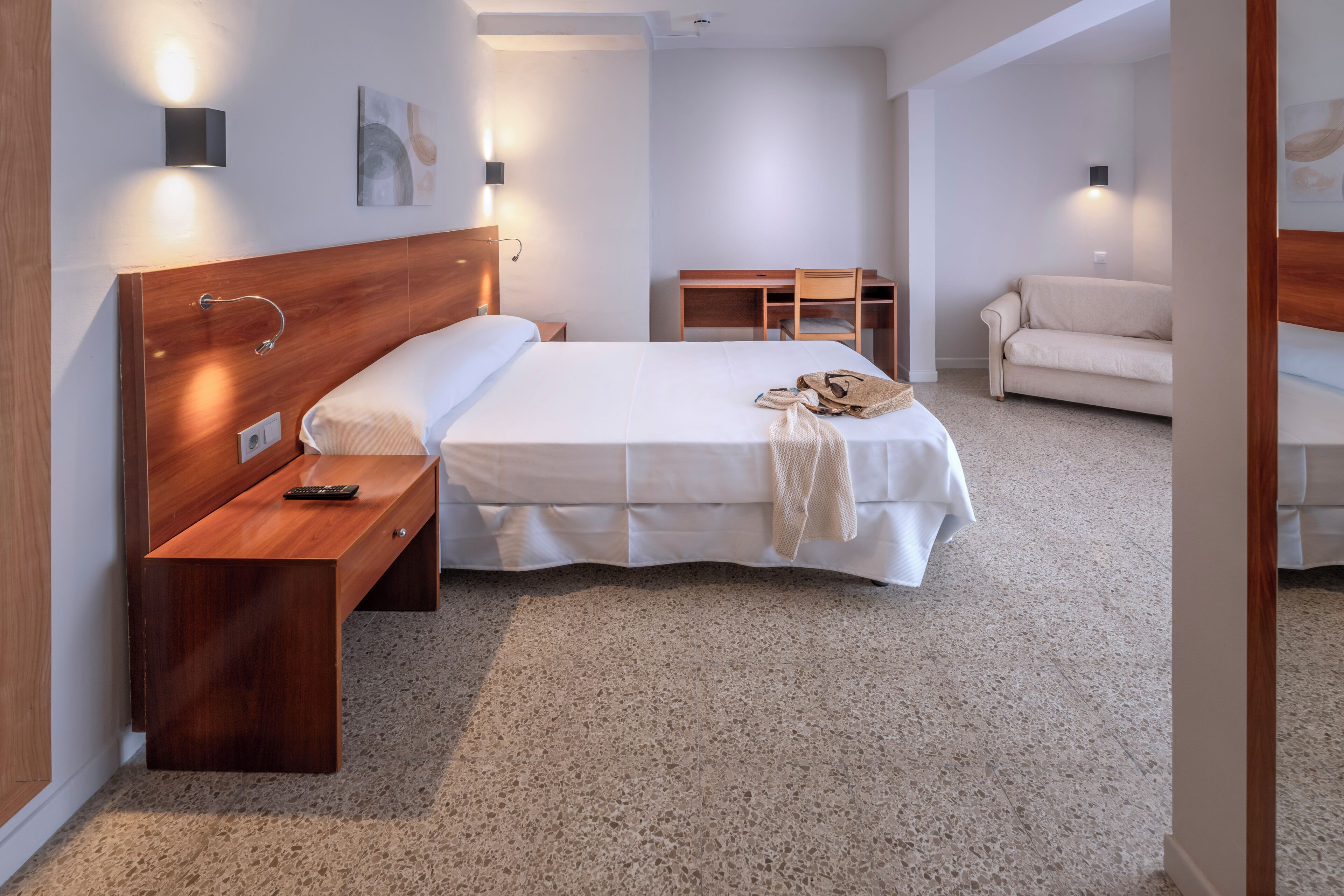 A hotel room with a wooden headboard, a bed with a white bedspread, a bedside table with a remote control, a wooden desk with a chair, and a sofa.
