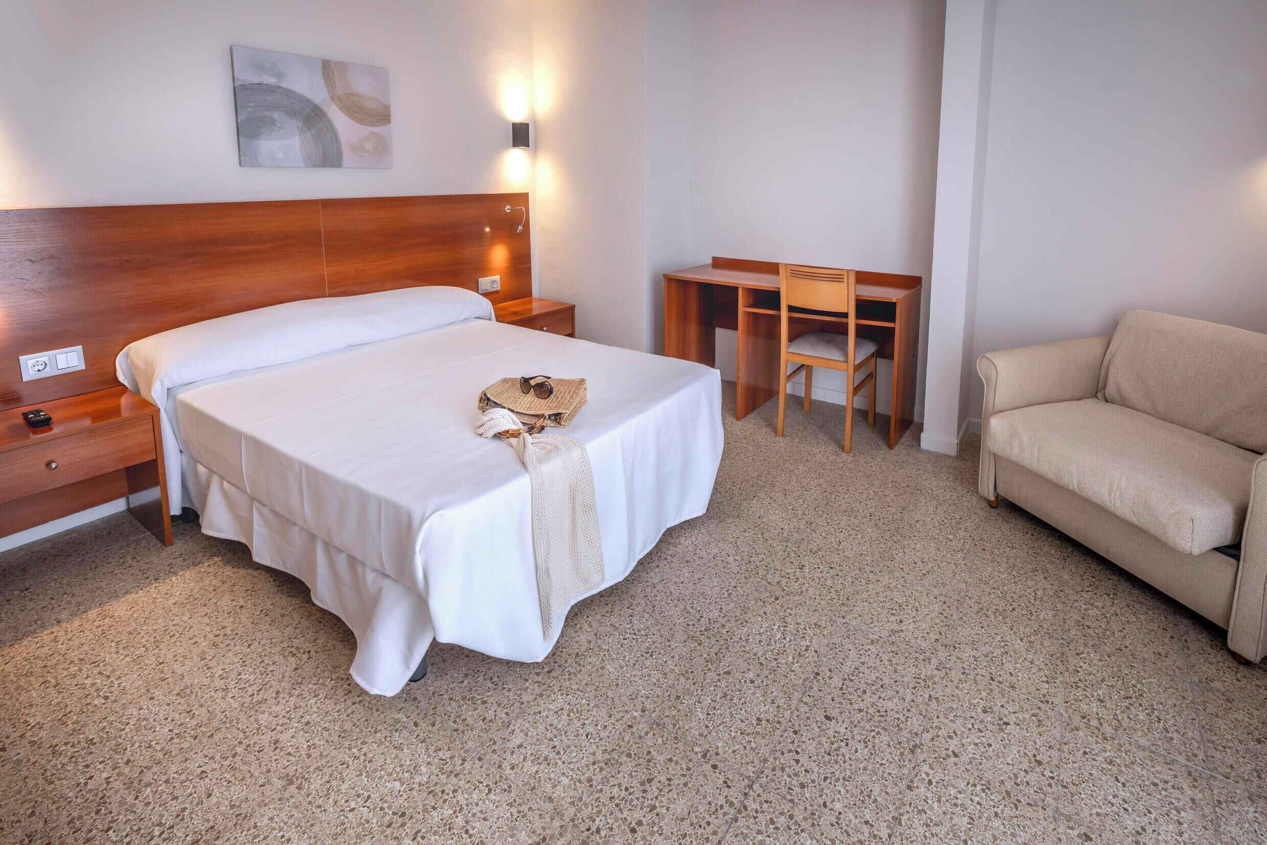 A hotel room with a bed, a desk, a chair, a sofa, and a wall-mounted light fixture.