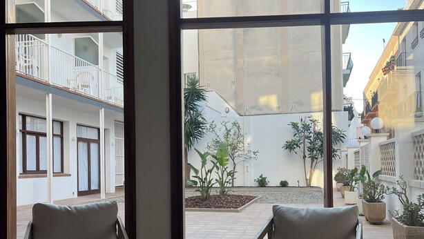 Ein Zimmer mit zwei Stühlen und einem kleinen Tisch, große Fenster mit Blick auf einen Innenhof und Gebäude.