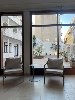 Una habitación con dos sillas y una mesita, ventanas amplias con vistas a un patio y edificios.