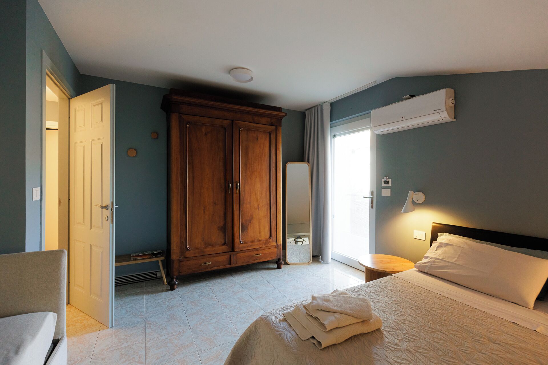 A hotel room with a bed, a wardrobe, a mirror, an air conditioning unit, and a bedside lamp.