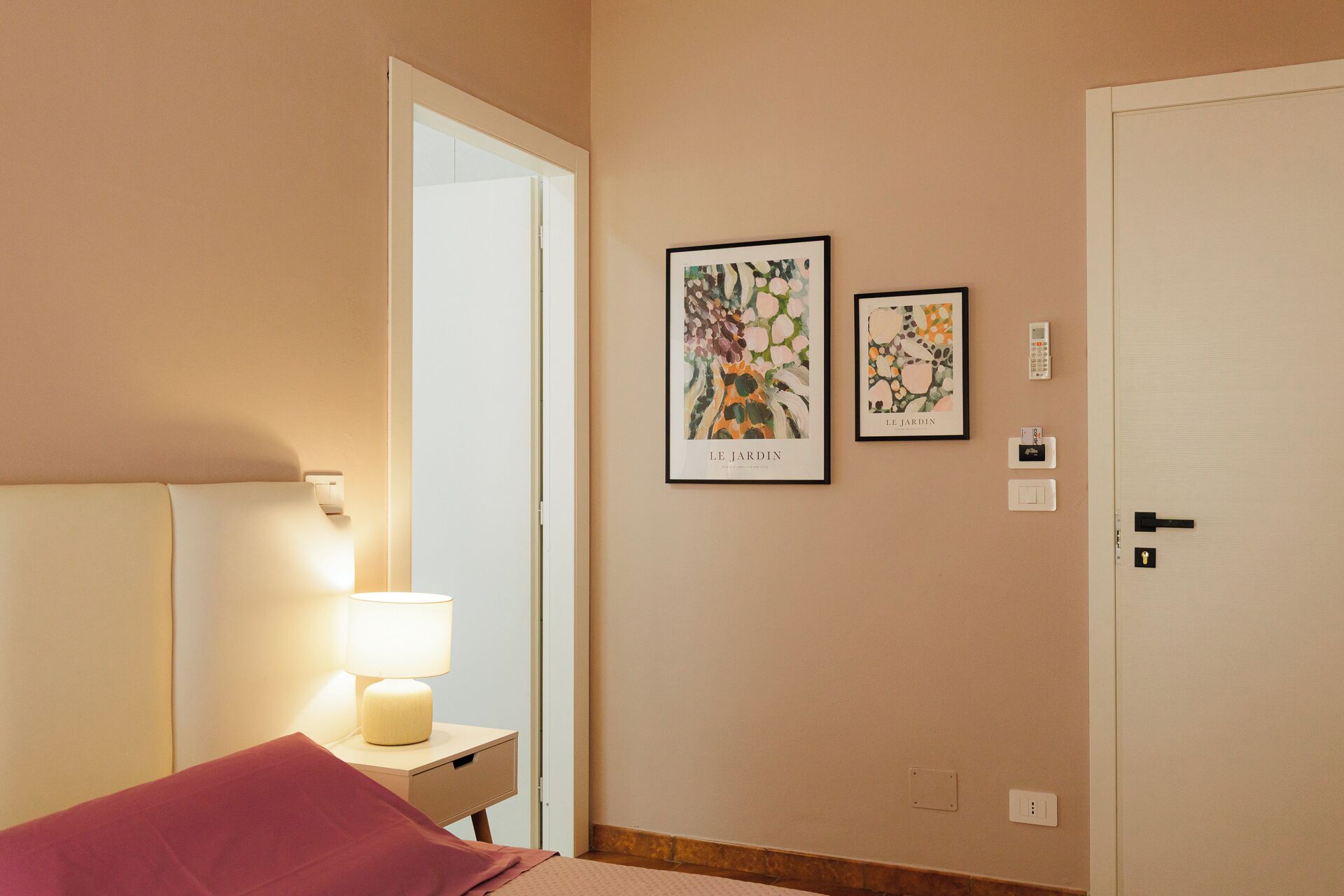 A hotel room with a bed, a bedside lamp, a door, and two framed artworks on the wall.