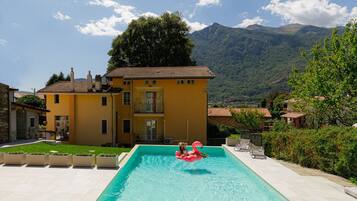 Ein Schwimmbecken mit zwei Personen, umgeben von einem Haus mit Garten und Bergen im Hintergrund.