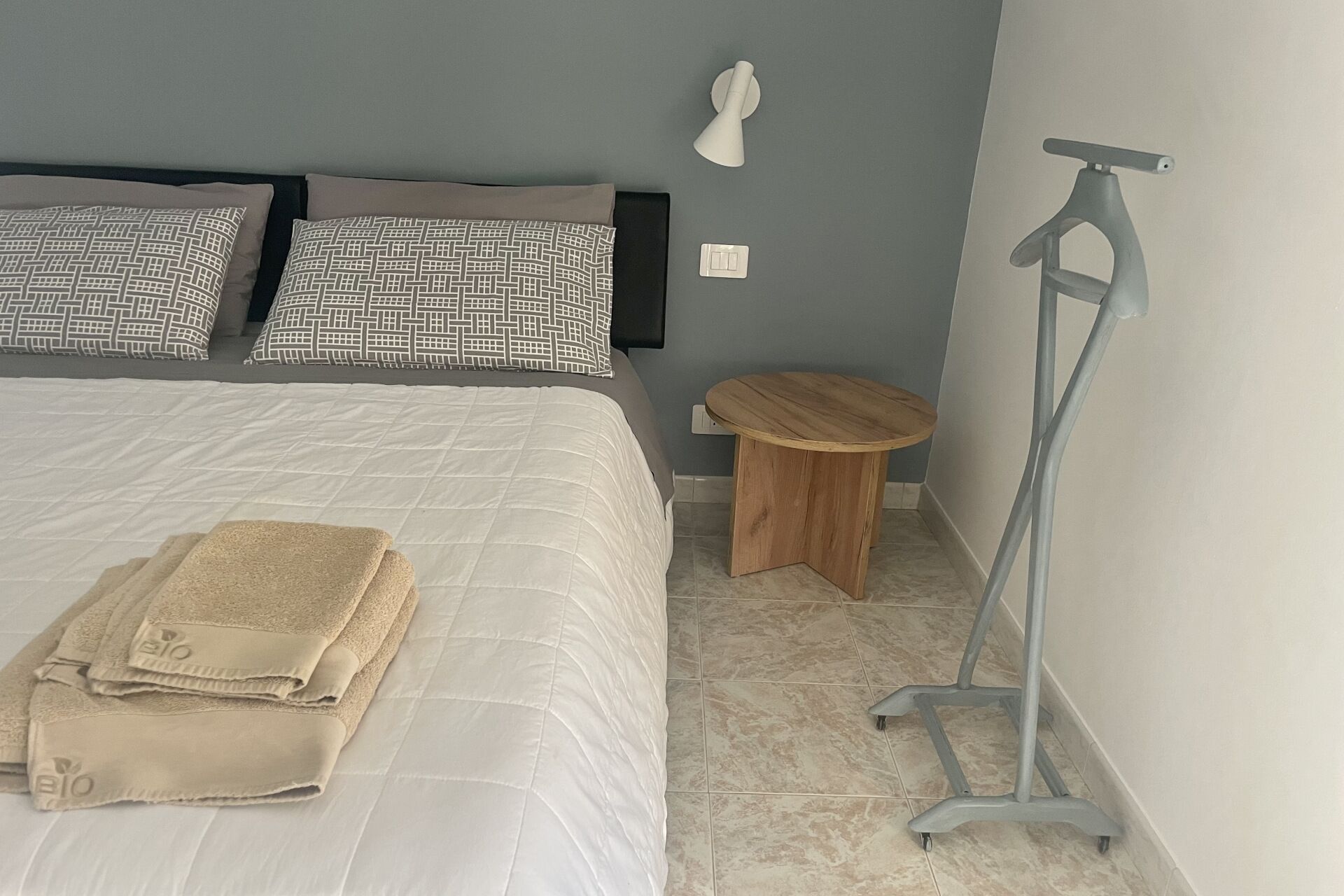 Uma cama bem arrumada com edredom branco e almofadas bege, um banquinho de madeira e um abajur.