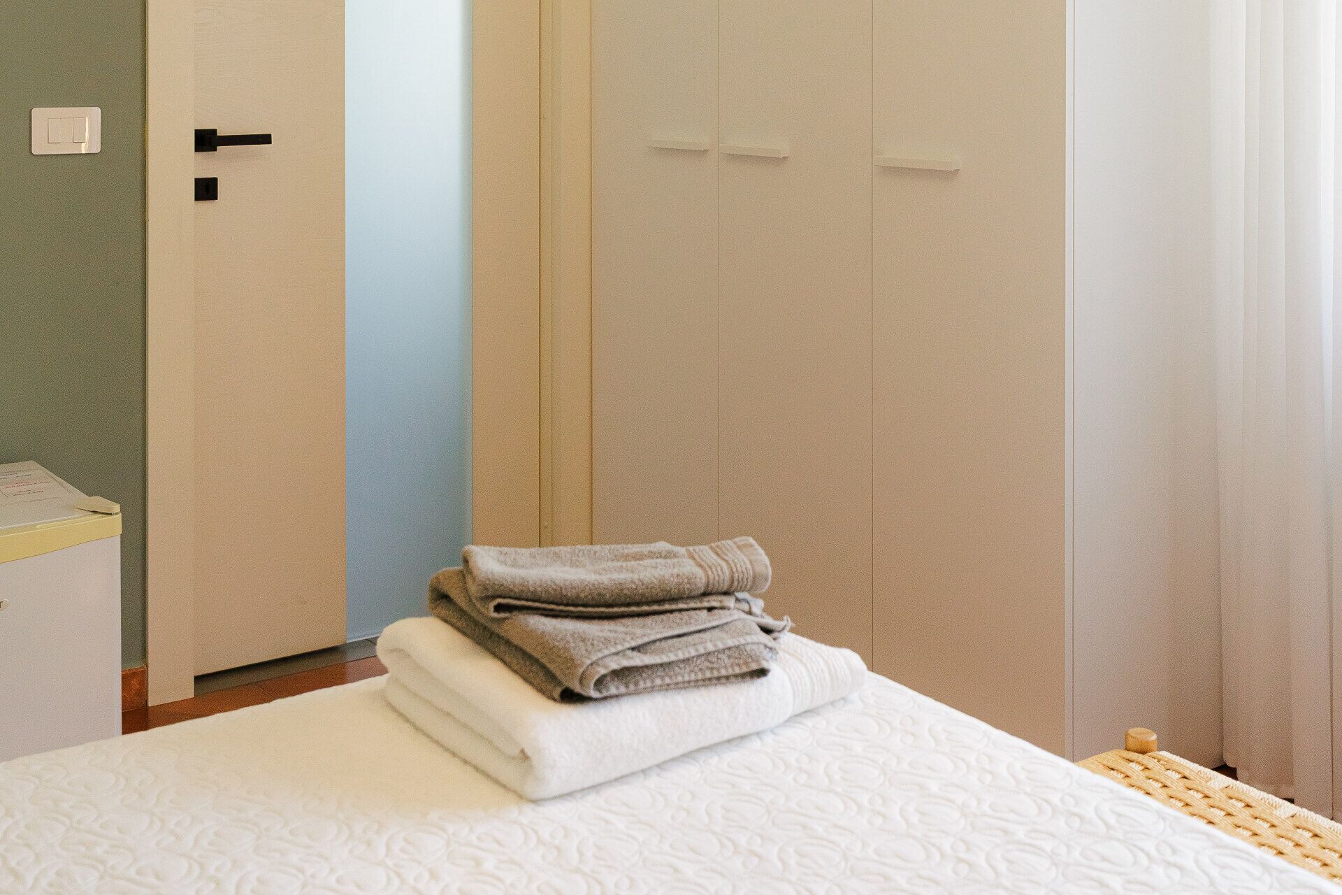 Una cama bien hecha con una pila de toallas dobladas, una mesita de noche de madera y una puerta con interruptor de luz.