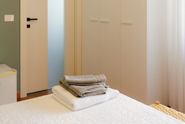 Una cama bien hecha con una pila de toallas dobladas, una mesita de noche de madera y una puerta con interruptor de luz.