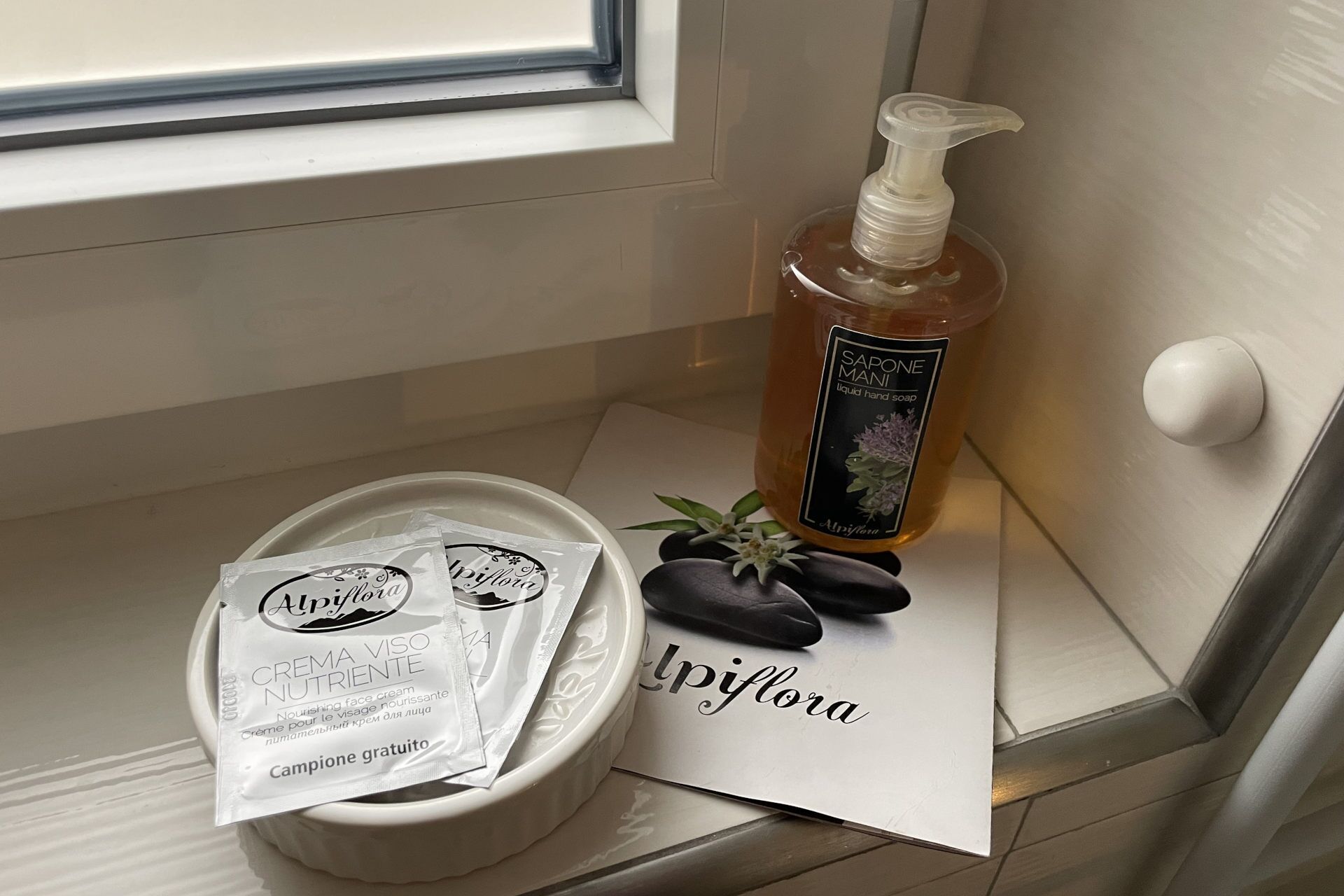 Un baño con ventana, una botella de jabón líquido, un paquete de crema facial y un objeto decorativo.