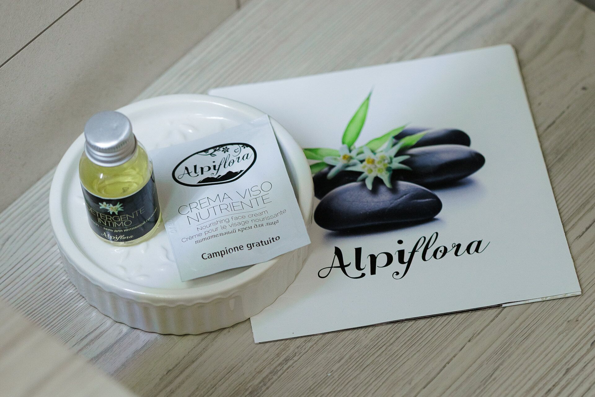Una bottiglia di crema viso nutriente Alpiﬂora, un contenitore di ceramica bianca e un depliant a tema spa.