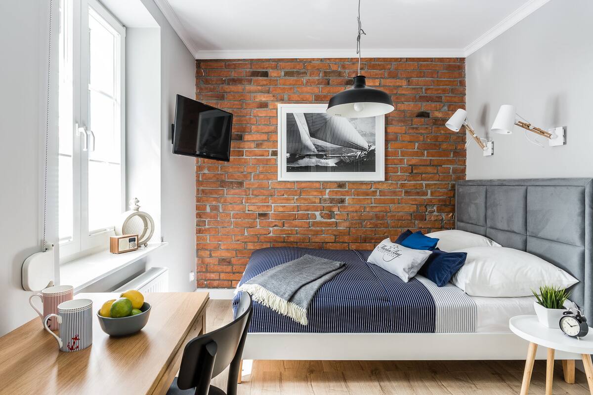 Un dormitorio moderno con pared de ladrillo, una cama con ropa de cama a rayas, cabecero gris, una mesita de noche con un cuenco de fruta, una silla, un televisor montado en la pared y una lámpara colgante.