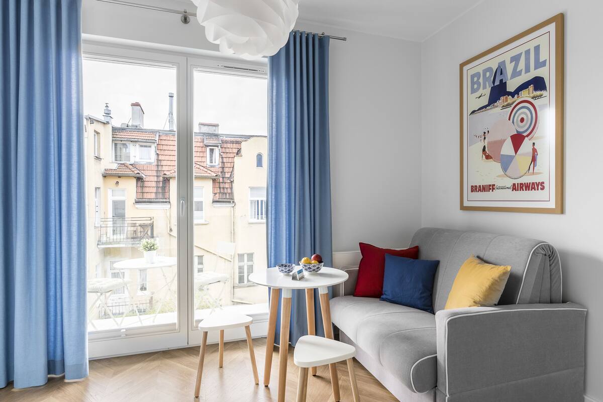 Una sala de estar moderna con un sofá gris, una mesita redonda con un frutero y un ventanal con cortinas azules.
