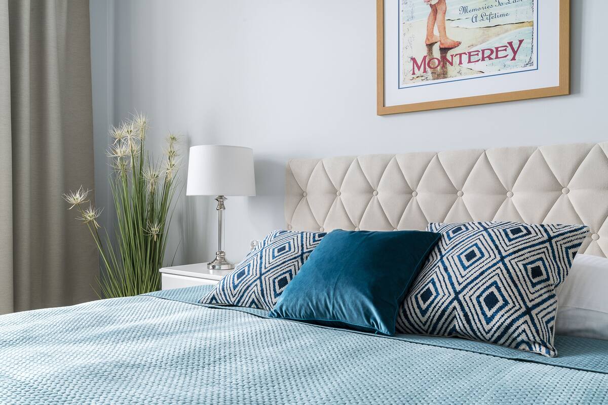 Un dormitorio con cabecera capitonada, cama con ropa de cama estampada, un cuadro en la pared y una lámpara de noche.
