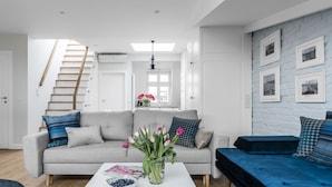 A modern living room with a grey sofa, a blue sofa, a coffee table with a vase of flowers, and a staircase leading to an upper level. - Sanhaus Apartments - Parkowa (Sopot)