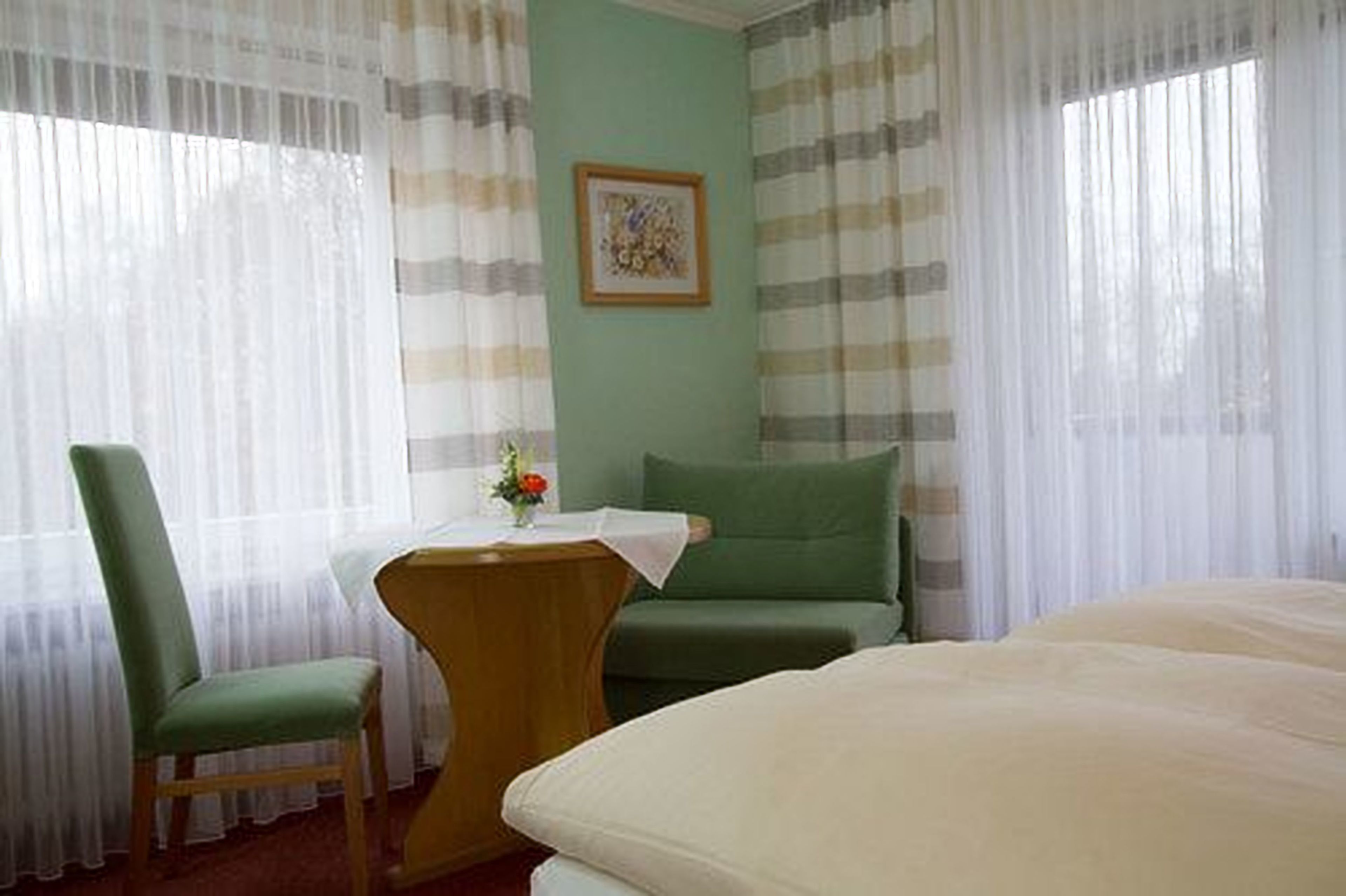 Une pièce comprenant un lit, une chaise, une petite table avec un vase de fleurs et un tableau accroché au mur.