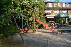 Un parque infantil con columpios y un tobogán, rodeado de árboles y un edificio al fondo.