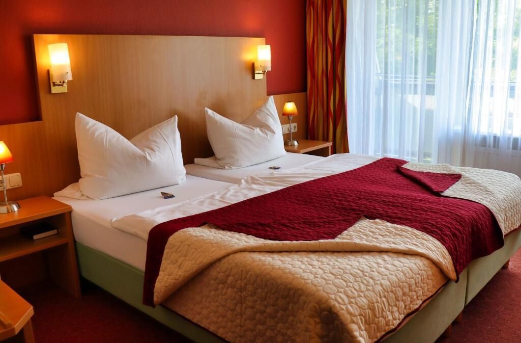 Ett hotellrum med två sängar, en sänggavel i trä, sänglampor och ett fönster med gardiner.
