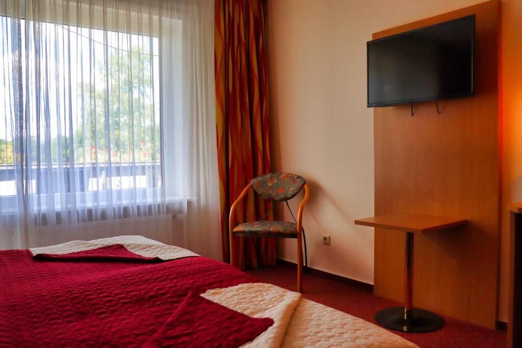 A hotel room with a bed, a chair, a TV mounted on the wall, and a window with curtains.
