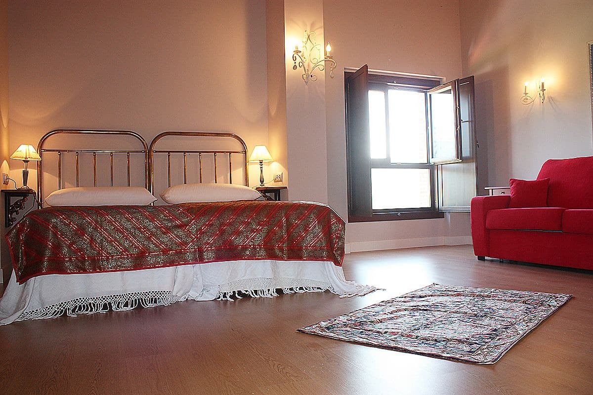 A bedroom with a bed, a red sofa, a window, and a wall-mounted lamp.