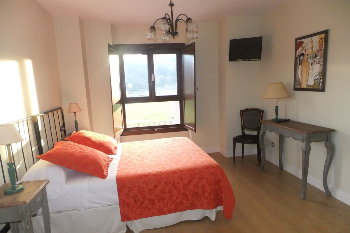A bedroom with a bed, a desk, a chair, a television, and a window with a view.