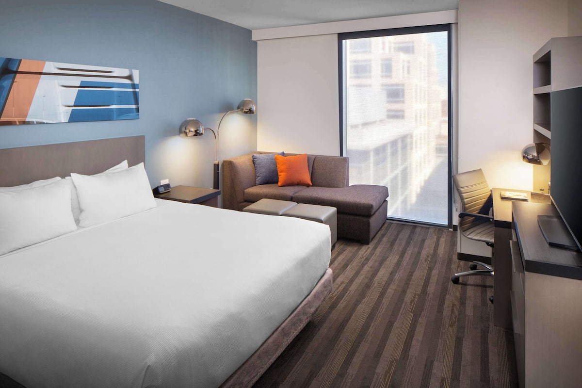 A hotel room with a large bed, a sofa, a desk with a chair, and a window with a city view.