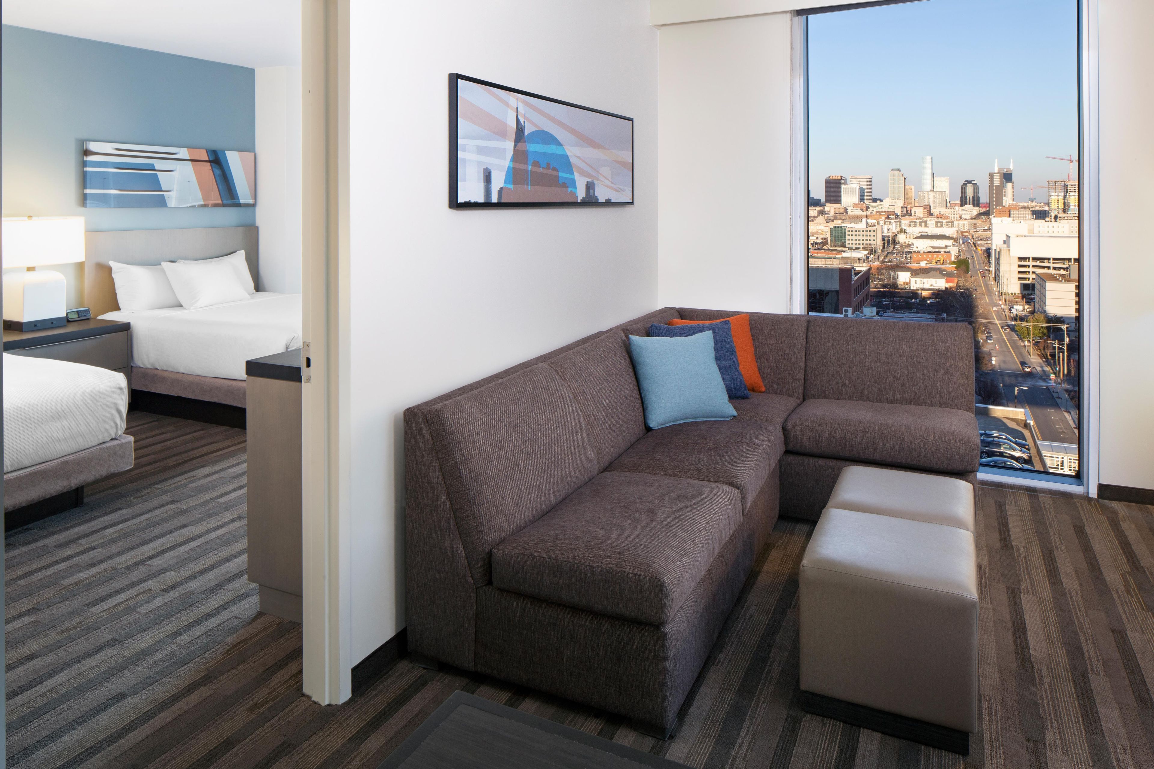 A modern hotel room with a large window offering a city view, a sectional sofa, a bed, and a small table.
