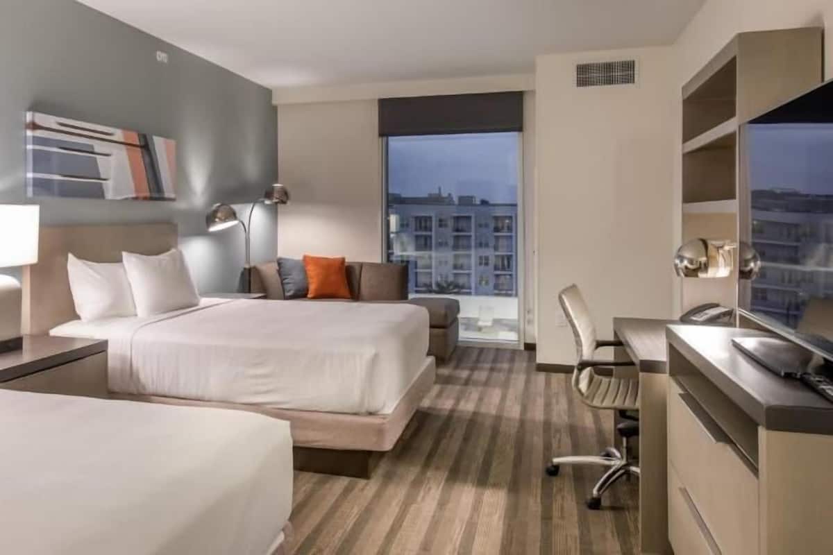 A hotel room with two beds, a desk, a chair, and a large window with a city view.