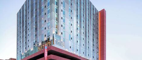 Een meerlaags hotel met een modern ontwerp, met een opvallende rode structuur aan de basis, grote ramen en een heldere lucht op de achtergrond.