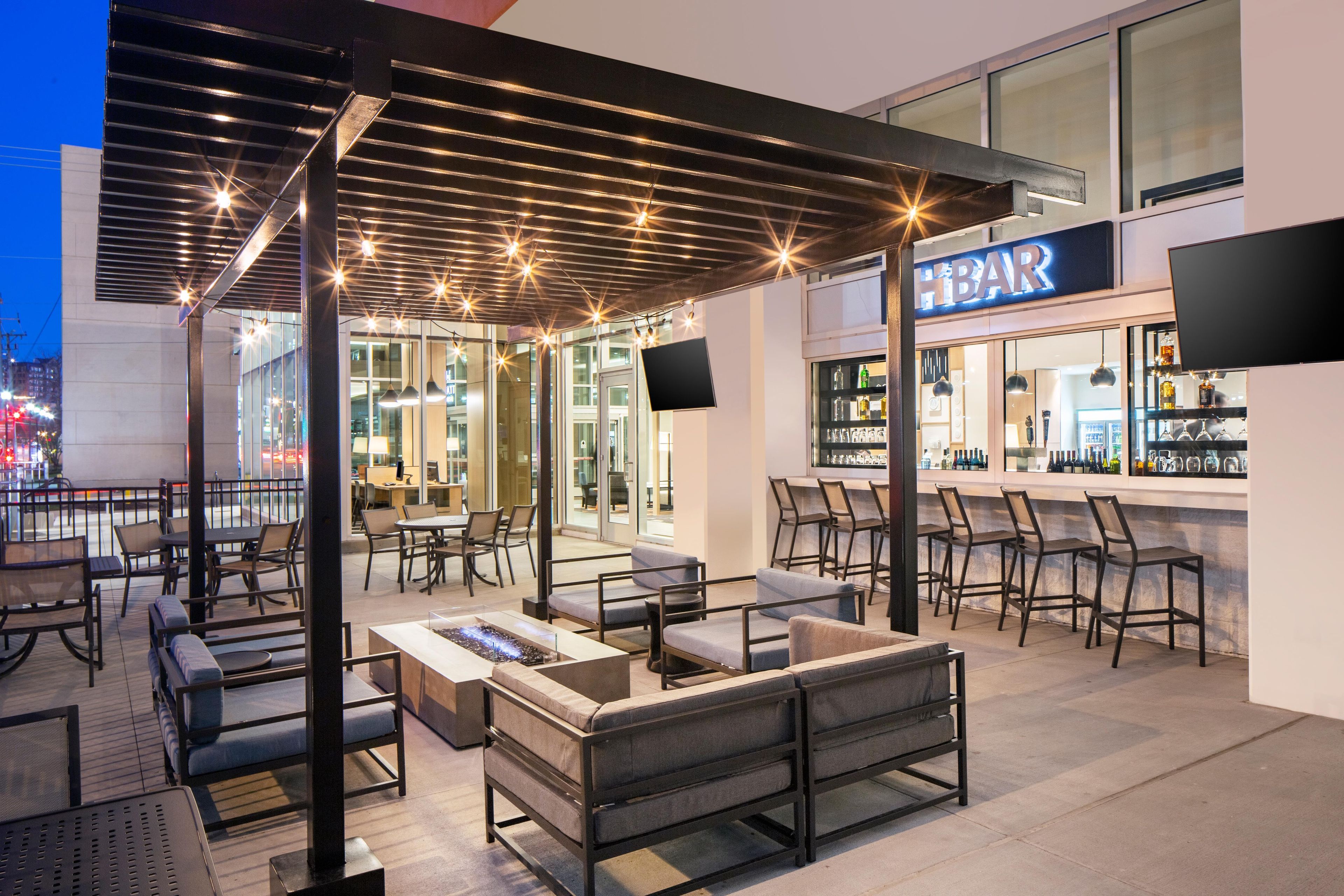 An outdoor seating area with tables and chairs, a bar with a visible sign, and a cityscape view in the background.