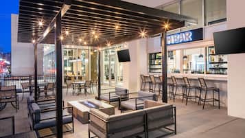An outdoor seating area with tables and chairs, a bar with a visible sign, and a cityscape view in the background.