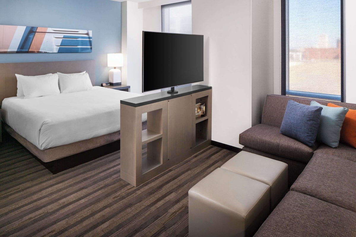 A hotel room with a bed, a TV on a stand, a sofa, and a window with a view.