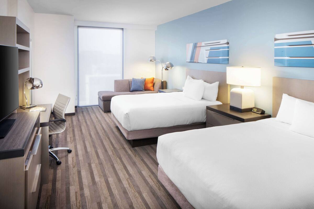 A hotel room with two beds, a desk, a chair, and a large window.
