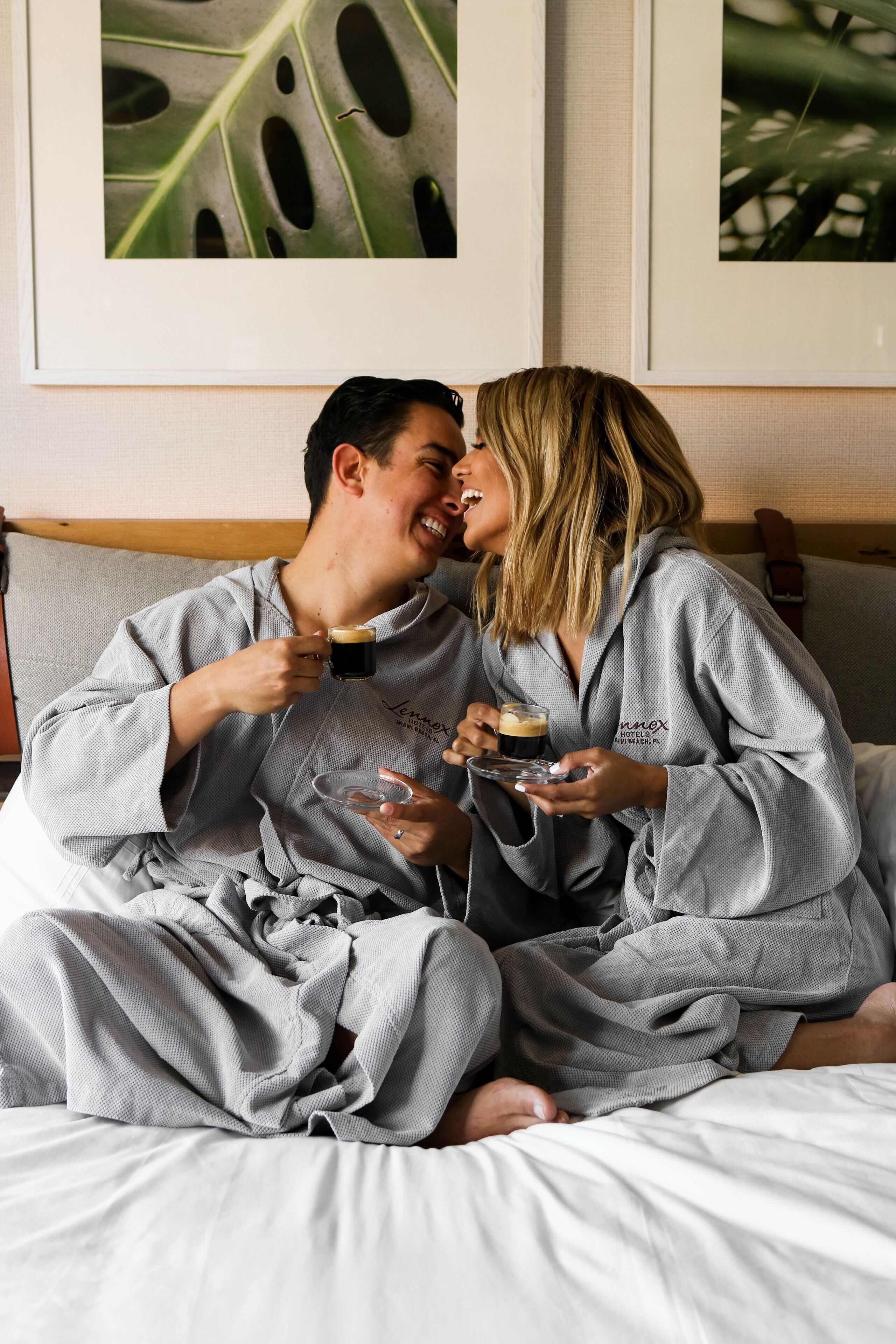 Una pareja con batas a juego sentada en una cama con cabecera de madera, sosteniendo vasos de una bebida oscura.