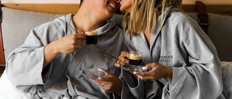 Una pareja con batas a juego sentada en una cama con cabecera de madera, sosteniendo vasos de una bebida oscura.