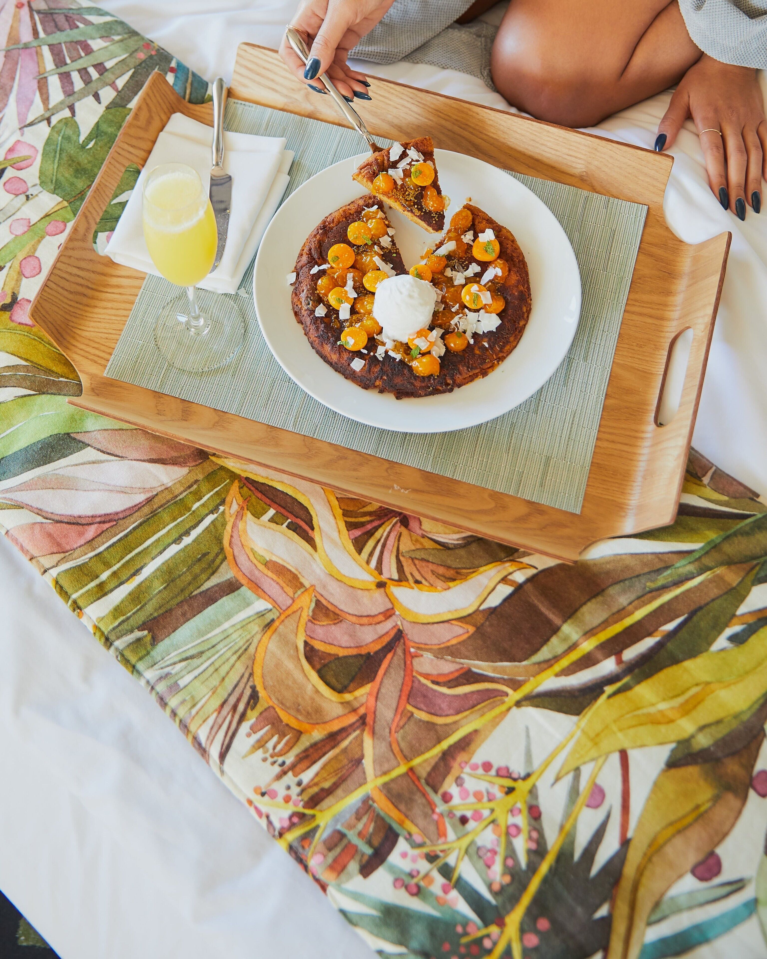 A breakfast tray with a plate of food, a glass of juice, and a fork on a bed with a colorful floral bedspread.