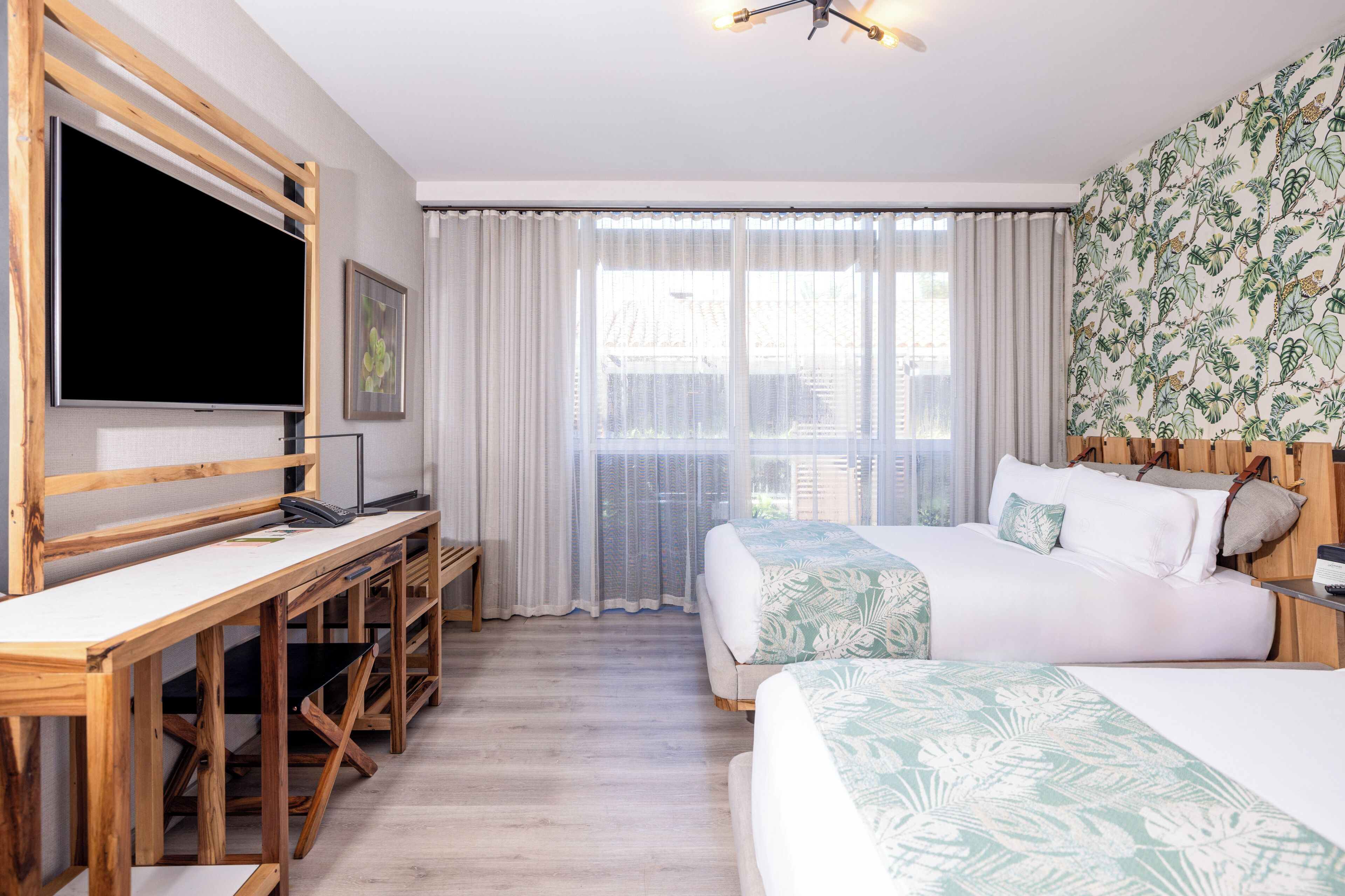 A hotel room with a large flat-screen TV, a desk with a chair, two beds with floral bedding, a balcony with a view, and a ceiling fan.