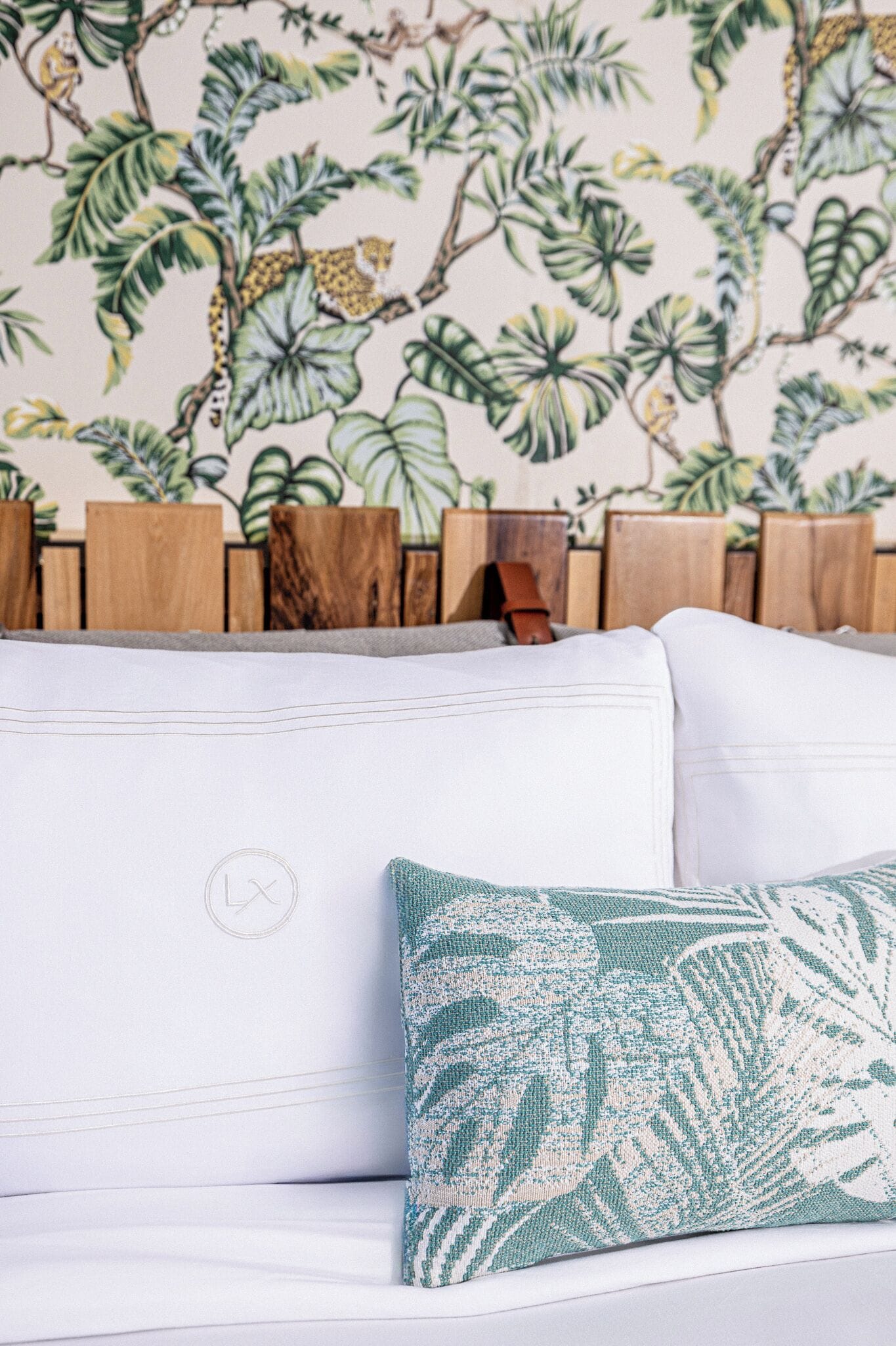 A white sofa with a green patterned pillow and a wooden headboard against a wall with a tropical leaf and fruit mural.