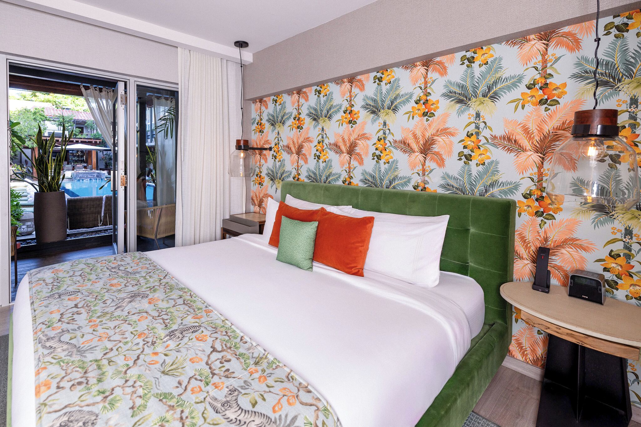 A hotel room with a large bed, a green headboard, a tropical wallpaper, a view of a pool, and a small table with a lamp.