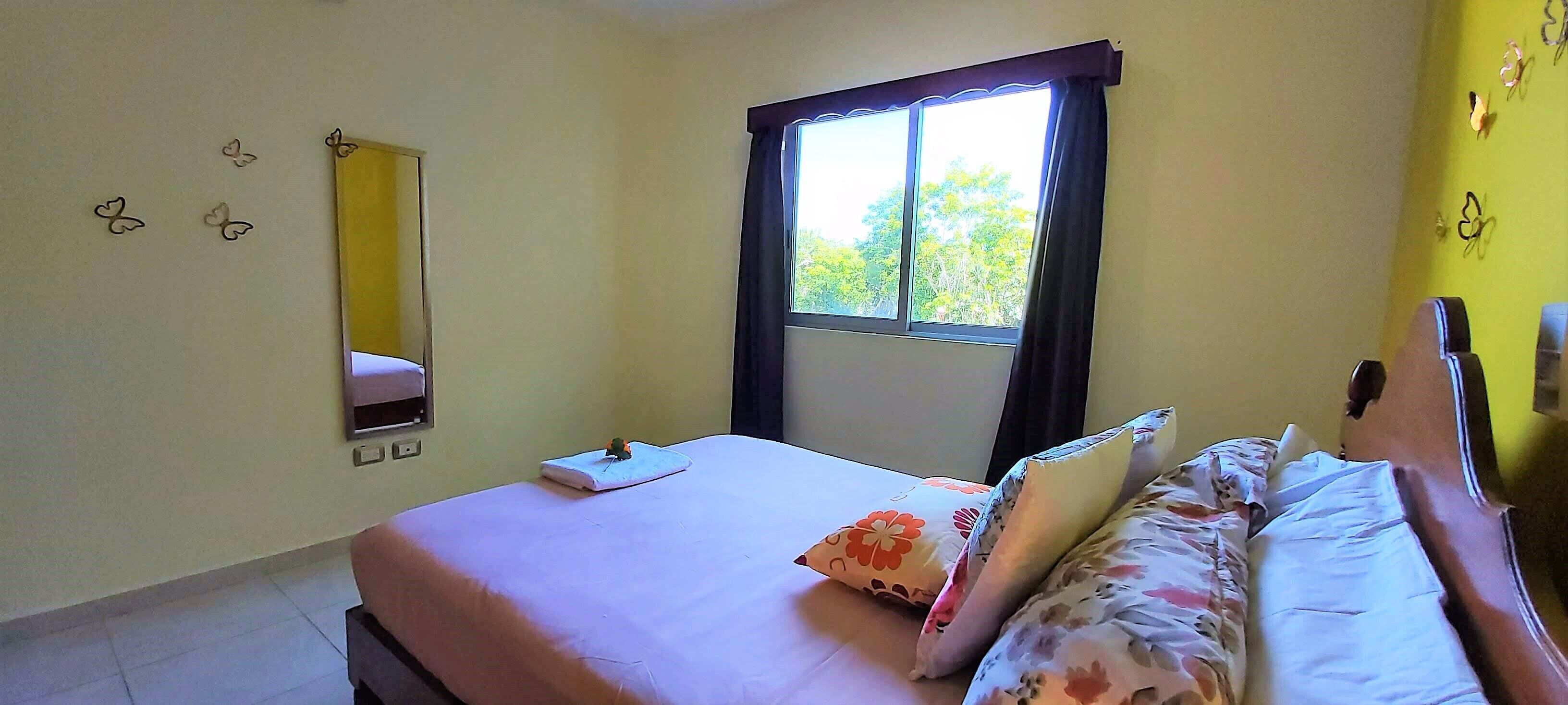 A bedroom with a bed, a window with curtains, a mirror on the wall, and a small plant on the bed.