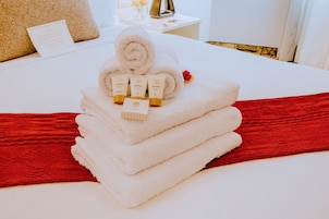 Una cama bien hecha con una colcha roja, una pila de toallas blancas y productos para el cuidado de la piel ZAYAH encima.