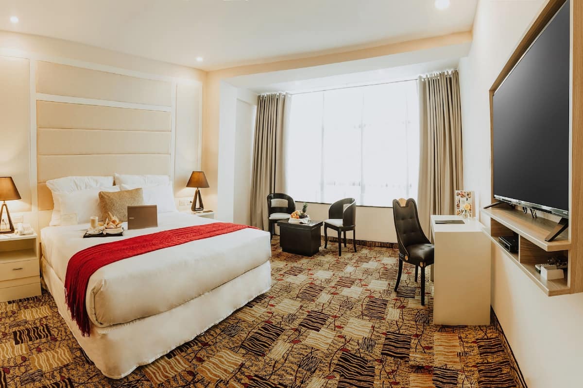 Quarto de hotel com uma cama grande, duas mesas de cabeceira com luminárias, uma escrivaninha com cadeira, televisão e uma janela com cortinas.