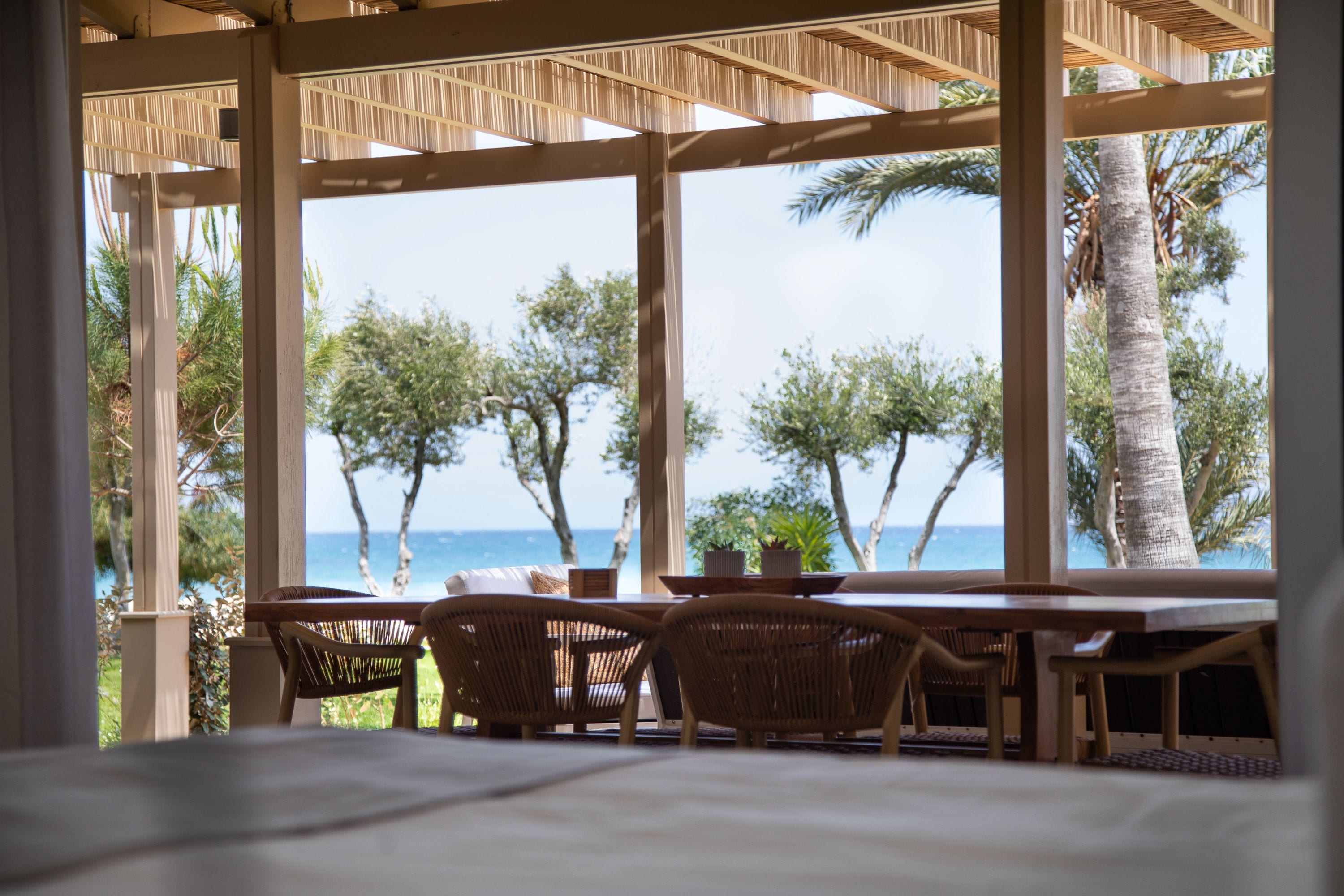 A covered outdoor seating area with a view of the ocean and palm trees.
