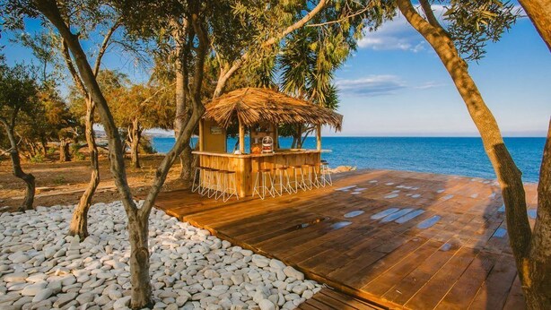 Una terraza de madera con una estructura de techo de paja, rodeada de árboles y piedras, con vista a un cuerpo de agua.