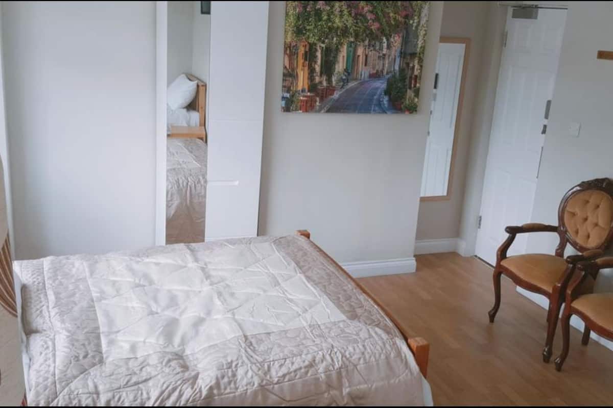 A bedroom with a bed, two wooden chairs, a painting of a street scene, and a white door.