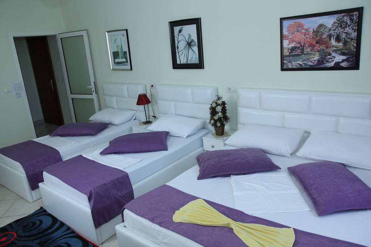 A hotel room with two beds, each with white bedding and purple pillows. There are framed pictures on the wall, a vase with flowers, and a red lamp on the nightstand.