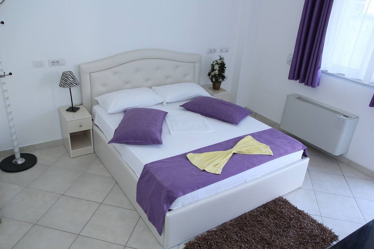 A neatly arranged bedroom with a bed, bedside tables, a lamp, a radiator, and a window with curtains.
