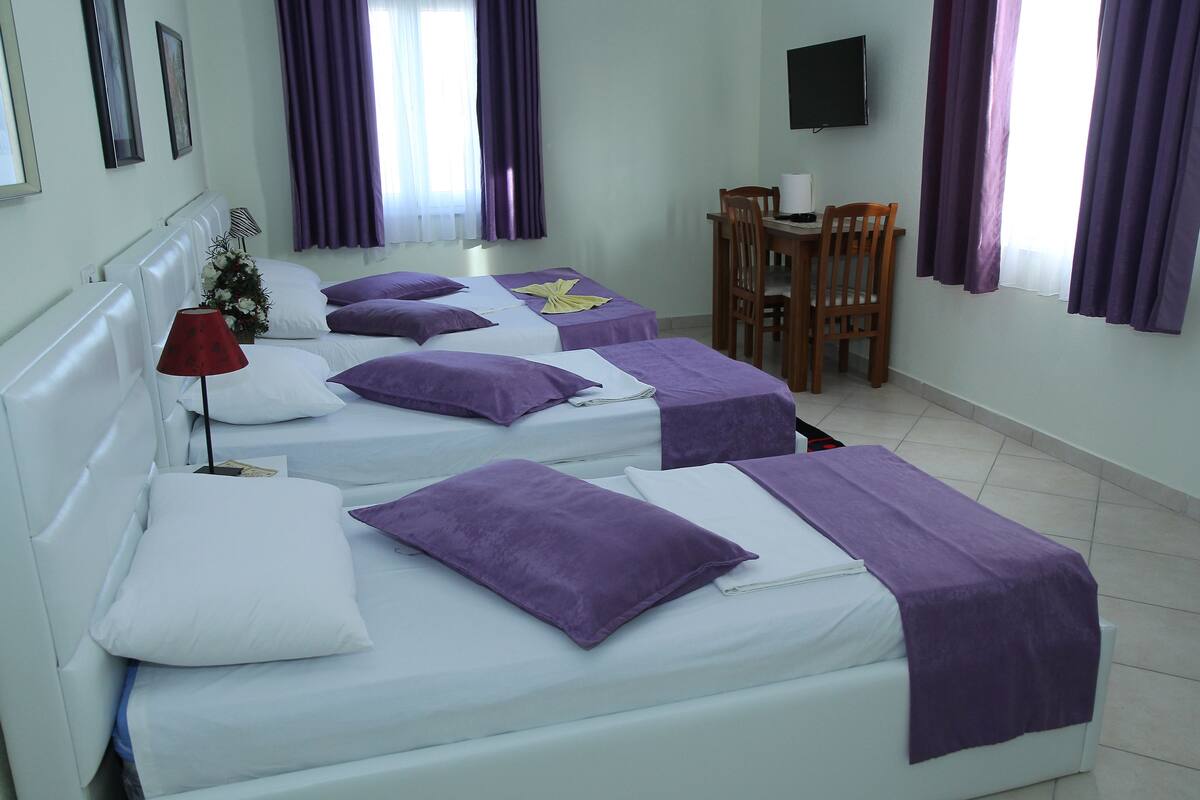 A hotel room with three beds, each with a purple blanket and white pillows. There is a desk with a chair, a TV mounted on the wall, and a window with purple curtains.