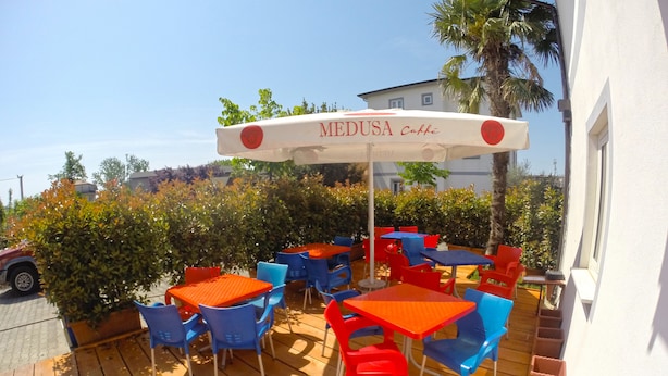 An outdoor patio with red and blue tables and chairs, a white umbrella with 'MEDUSA Caffè' written on it, and a white building in the background. - Hotel Bicaj (Shkodër)