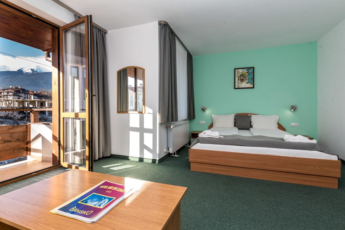 A hotel room with a bed, a table with a magazine, a view of mountains, and a window with curtains.